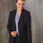 A person wearing a black blazer and grey shirt smiles while standing in front of a grey textured wall.