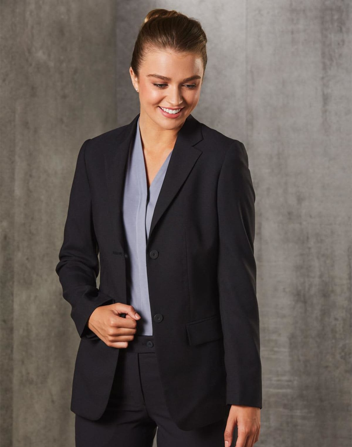 A person wearing a black blazer and grey shirt smiles while standing in front of a grey textured wall.