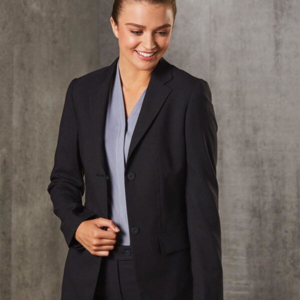 A person with tied-back hair wears a black blazer over a gray shirt, smiling slightly while looking down. The background is a textured gray wall.