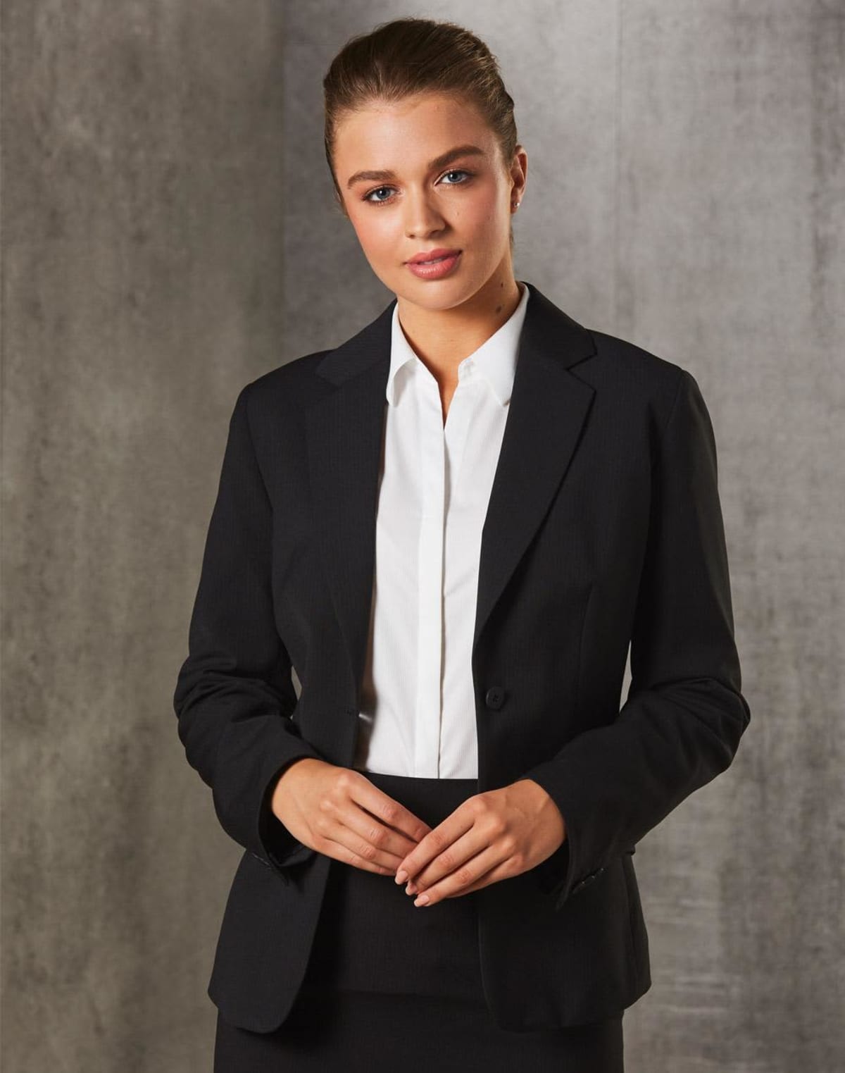A person wearing a black blazer and white shirt, standing with their hands clasped in front. The background is a textured gray surface. The person has a neutral expression and neatly styled hair.