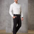 A man stands confidently wearing a white dress shirt and black pants, with hands in pockets. He has a neat hairstyle and is posing against a textured gray wall.