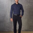 A man in a dark blue dress shirt and black pants stands against a gray textured background, looking at the camera. He is wearing black shoes and a black belt.