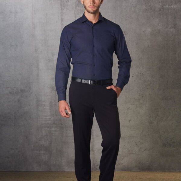 A man stands against a textured gray wall, wearing a dark blue dress shirt, black trousers, and black shoes. His hands are by his sides, and he looks directly at the camera.