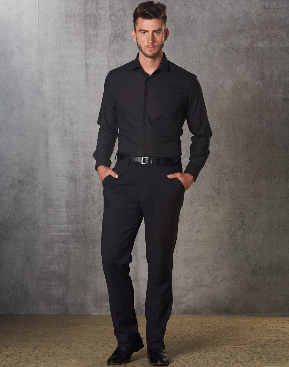 Man in a dark shirt and pants stands against a textured gray wall.