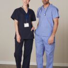 Two medical professionals stand together. One is wearing dark scrubs with an ID badge, and the other is in light blue scrubs with a stethoscope around their neck. Both are wearing white sneakers and standing against a neutral background.