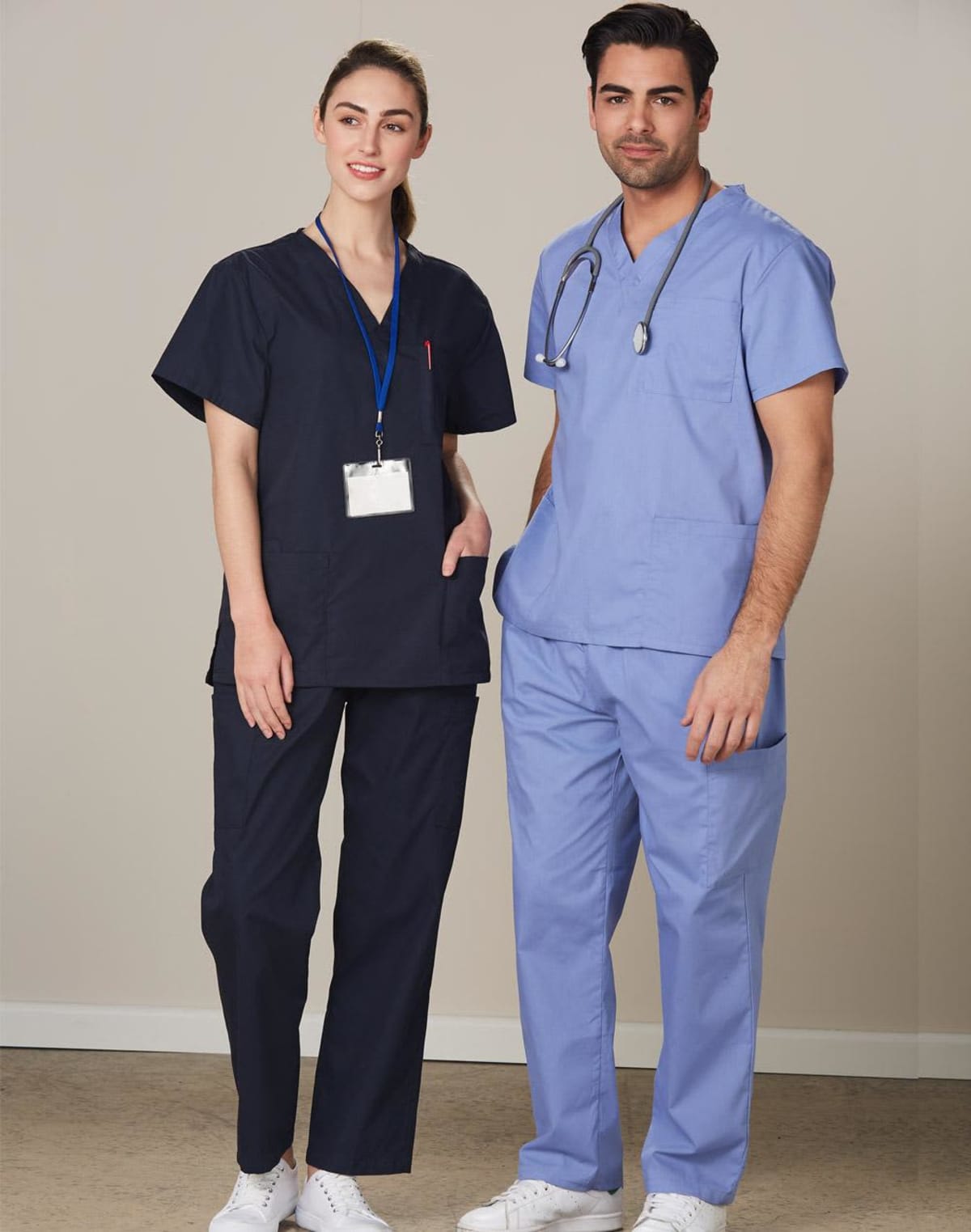 Two medical professionals stand together. One is wearing dark scrubs with an ID badge, and the other is in light blue scrubs with a stethoscope around their neck. Both are wearing white sneakers and standing against a neutral background.