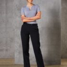 A person stands with arms crossed, wearing a light grey blouse and black trousers, against a concrete wall background. The individual has short, blonde hair and is wearing black heels.