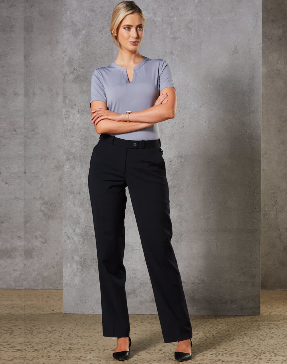 A person stands with arms crossed, wearing a light grey blouse and black trousers, against a concrete wall background. The individual has short, blonde hair and is wearing black heels.