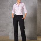 A person stands against a gray textured wall, wearing a light pink button-up shirt and black dress pants. They have their hands in their pockets and are wearing black shoes.