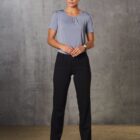 A person stands against a textured gray wall, wearing a light gray short-sleeve top and dark pants. They have their hair tied back and are wearing black shoes. Their hands are loosely clasped in front.