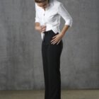 A woman in a white shirt and black pants adjusts her waistband while looking down. She stands against a textured gray wall, with her hair tied back and wearing black shoes.
