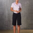 A person stands against a gray concrete wall wearing a light pink short-sleeved shirt, dark knee-length shorts, and black shoes. They have their hair tied back and are looking forward with a neutral expression.