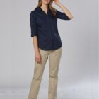 A person with long hair is wearing a navy blue button-up shirt and beige pants, standing on a plain light-colored background. They have one hand in their pocket and are smiling while touching their hair.