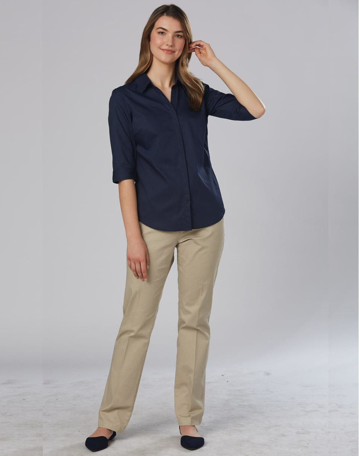 A person with long hair is wearing a navy blue button-up shirt and beige pants, standing on a plain light-colored background. They have one hand in their pocket and are smiling while touching their hair.