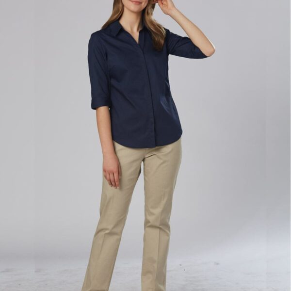 A person with long hair is standing against a plain gray background, wearing a dark blue button-up shirt, beige pants, and black shoes. They are smiling and have one hand raised near their head.