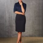 A woman stands confidently in front of a textured gray background. Shes wearing a navy blouse with sleeves rolled up and a knee-length black skirt. Her arms are crossed and she wears black heels, smiling softly.