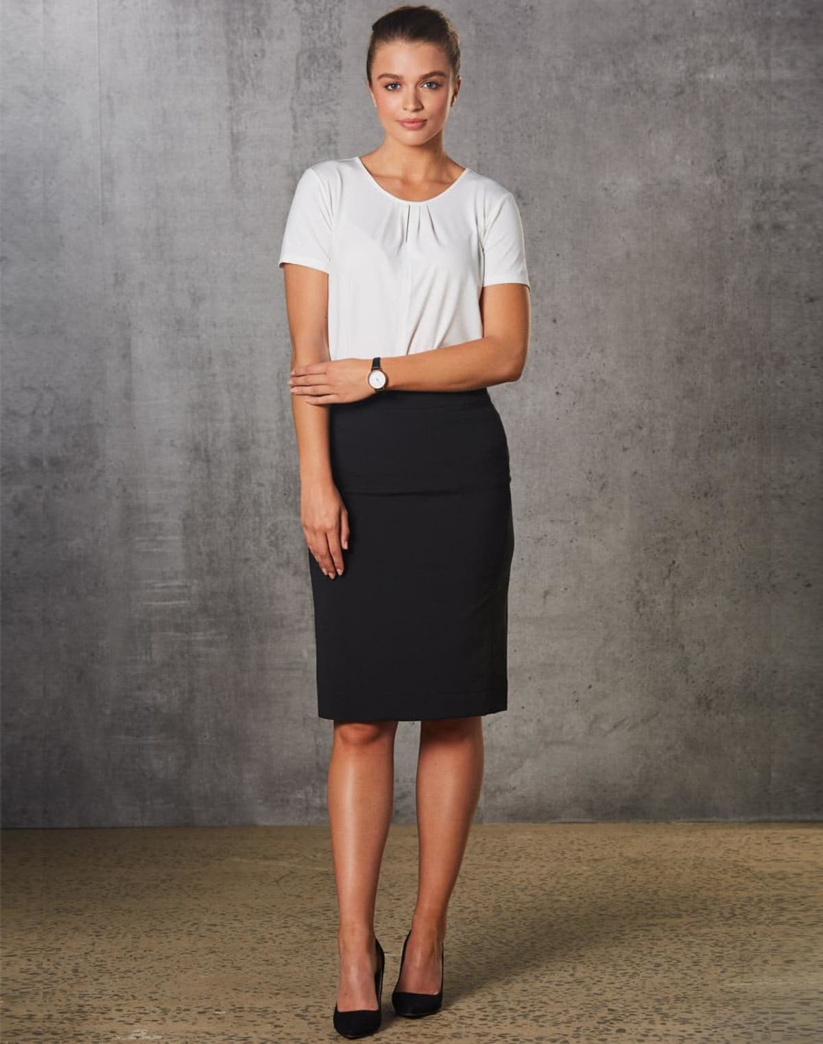 A woman stands against a gray textured wall. She wears a white shirt, black skirt, black high heels, and a watch on her left wrist. Her hair is neatly tied back, and she stands with her arms gently crossed.