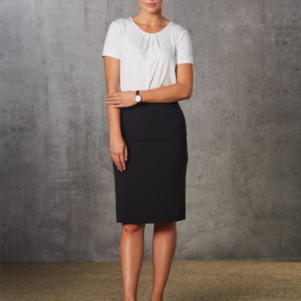 A woman stands against a textured gray wall, wearing a white blouse and a black knee-length skirt. She has her arms crossed and is wearing black shoes and a watch, with hair styled in a bun.