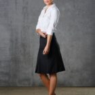 A person stands against a gray textured wall, wearing a white blouse, black knee-length skirt, and black shoes. They have one hand on their hip and the other at their side, looking towards the camera with a neutral expression.