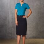 A person stands in front of a gray concrete wall, wearing a teal blouse, a dark knee-length skirt, and black high heels. Their hair is styled up, and they are looking forward with a slight smile.