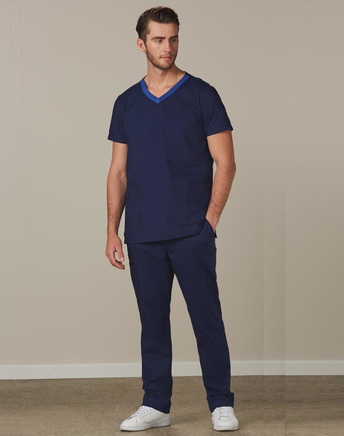 Man wearing navy blue scrubs and white sneakers standing against a plain beige wall.