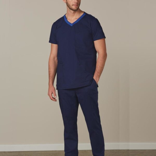 Man wearing navy blue medical scrubs and white sneakers, standing indoors against a plain wall.