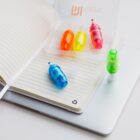 Colored correction tape pens in blue, orange, pink, yellow, and green are displayed on an open lined notebook resting on a closed laptop. A transparent plastic case is shown, partially open, containing the pens.