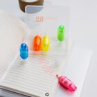 Image of a spiral notebook open on a desk with a pink squiggle drawn on the page. Nearby are four highlighters in blue, yellow, green, and pink, standing upright. They are stored in a transparent case labeled with Imoxi.