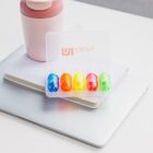 A case of five colorful push pins is open on top of a laptop and a closed notebook. A pink, lidded tumbler is placed beside the case. The scene is set on a bright surface.