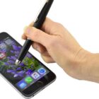 A hand uses a stylus on a smartphone with a purple flower wallpaper. The screen displays various app icons.