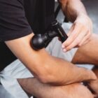 A person using a black Theragun-like massage device on their arm, seated on a chair. They are wearing a black t-shirt and light-colored shorts. The device has RECKMAH  inscribed on it.