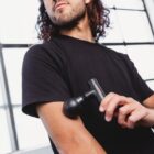 A person with long hair uses a black handheld massage gun on their upper arm. They are wearing a black T-shirt and standing in front of a large window with natural light streaming in.