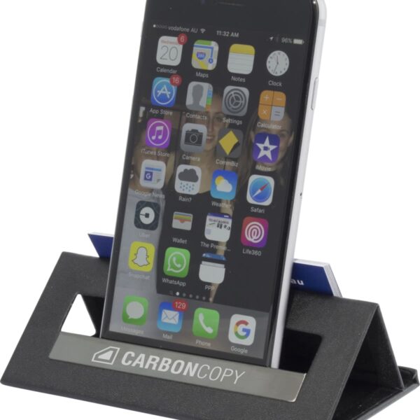 A smartphone displaying app icons rests on a black carbon fiber stand labeled CARBONCOPY. The stand has a slot holding business cards behind the phone.