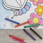 Coloring page featuring a butterfly and flowers next to scattered crayons in orange, green, blue, violet, red, and black. The butterfly is partially colored, lying on a wooden surface.