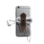 A smartphone with attached white earphones is secured with a brown strap featuring a cow illustration and text. The strap neatly organizes the earphone cables, resting on the back of the phone.