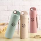 Three insulated water bottles with handles, each in pastel colors: green, beige, and pink, are displayed on a light wooden surface. The bottles have a circular emblem with CUP printed on them. A wicker basket and a white brick wall are in the background.