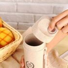 A person opens a beige insulated cup with a matching cap. The cup has a logo and is on a table with a wicker basket containing fruits, such as oranges and apples. The background features a white brick wall.
