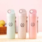 Three insulated bottles with handles are lined up against a pink background. They are pastel green, beige, and pink, each with the text CUP and a laurel wreath design. A wicker basket with fruits is blurred in the background.