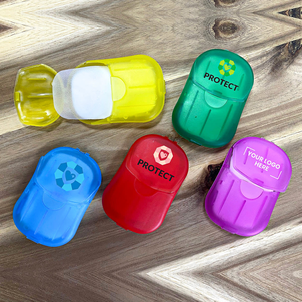 Colorful plastic cases containing sheets of paper soap are spread on a wooden surface. The cases are yellow, green, red, purple, and blue, each labeled with PROTECT and a space for a logo or design.