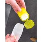 A person holding a translucent soap sheet in one hand and a small, open, yellow plastic case in the other. The case has a single soap sheet inside. The background shows a dark gray carpeted floor.