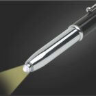 A close-up of a sleek black pen with a built-in LED flashlight at the tip, casting a yellow beam of light against a dark gray background.