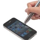 A hand holding a stylus is interacting with an iPhone, touching its screen. The phone displays various app icons on its home screen. The image focuses on the action of using the stylus with the smartphone.