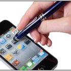 A hand using a stylus pen on a smartphone screen displaying various app icons. The background is plain white.