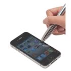 A hand using a stylus to tap on the screen of a smartphone displaying various app icons. The phone is placed on a white surface.