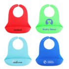 Four colorful silicone bibs are displayed. The top row shows a red bib with Sovamoo and a green bib with Baby Bear. The bottom row shows a light blue bib with Idero and a blue bib with Foroda Company. Each has adjustable neck fasteners.