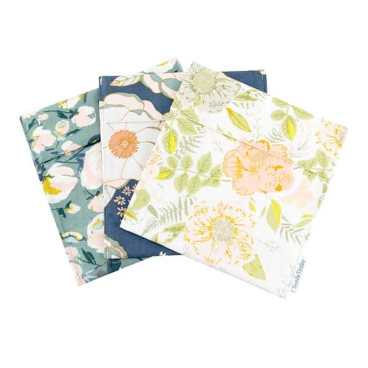 Three reusable snack bags are displayed, featuring floral patterns. One bag has a dark background with light flowers, another has a light beige design with colorful flowers, and the third showcases yellow and green floral patterns on a white background.