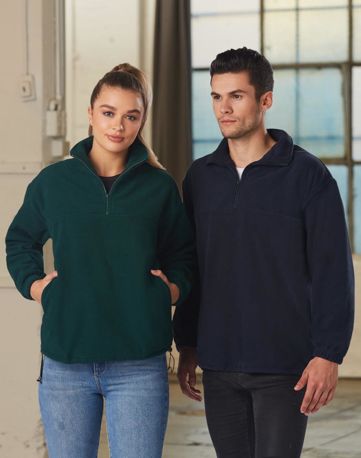 A woman and a man standing side by side indoors. The woman is wearing a green half-zip pullover and jeans, while the man is wearing a black half-zip pullover. Both are looking at the camera with neutral expressions.