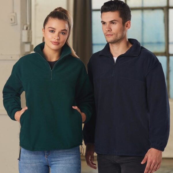 A woman and a man stand side by side indoors. The woman wears a dark green pullover and jeans, and the man wears a navy pullover with matching pants. Both have their hands in their pockets and are looking forward.
