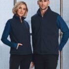 Two people, one with short blonde hair and the other with short brown hair, are wearing matching navy blue vests over long-sleeve blue shirts. They are standing in front of a large window with a bright background.
