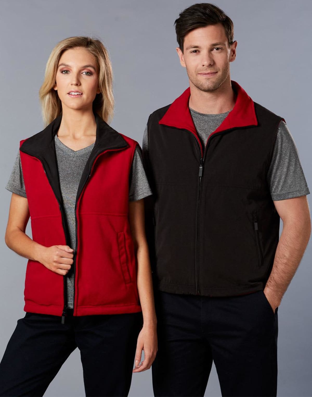 A woman and a man stand side by side, both wearing gray t-shirts and zip-up vests. The woman’s vest is red with black trim, and the mans vest is black with a red collar. They are looking at the camera against a plain gray background.